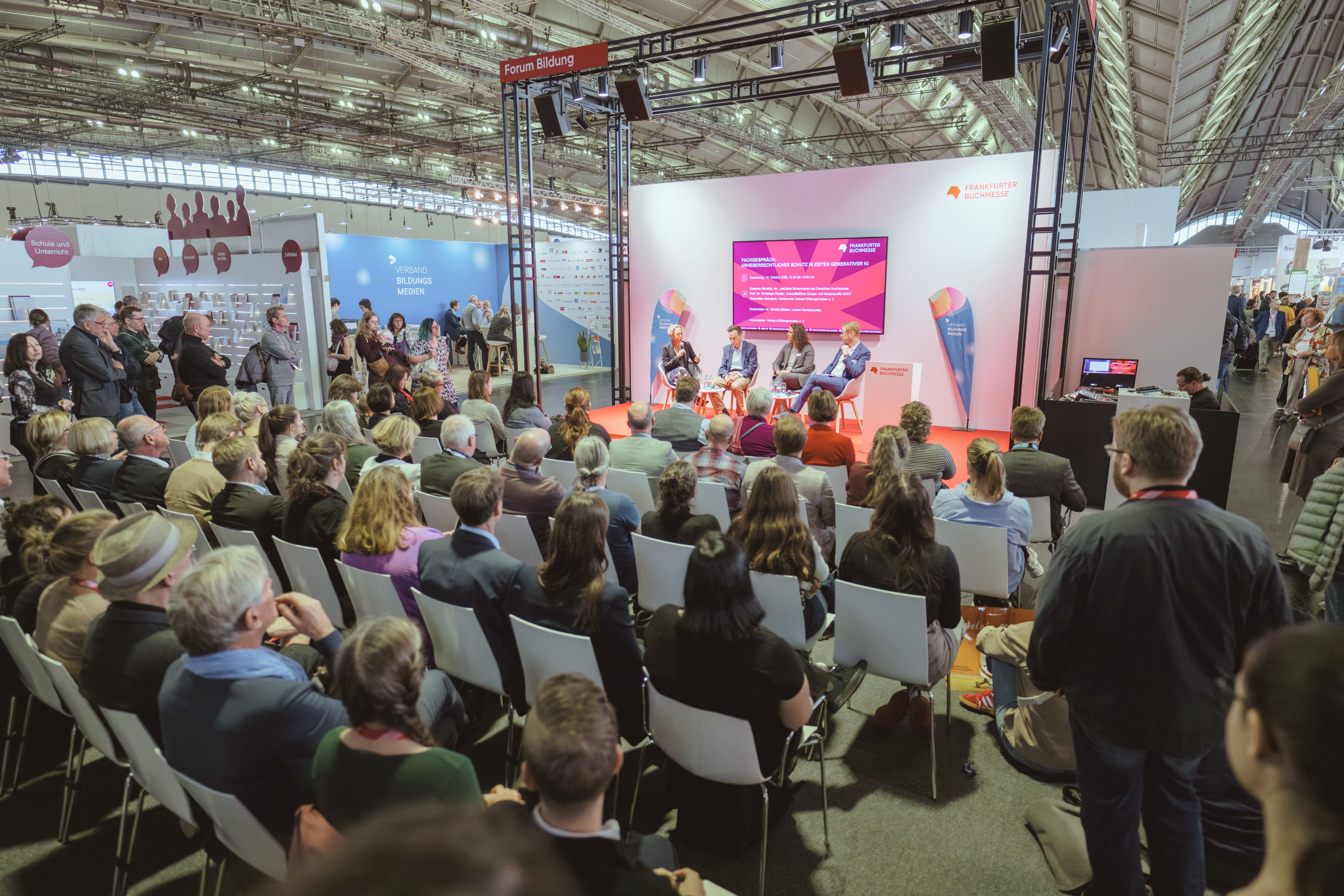 Zuschauerinnen und Zuschauer sitzen in einer Messehalle auf Stuhlreihen und schauen auf eine Bühne, wo eine Podiumsdiskussion stattfindet